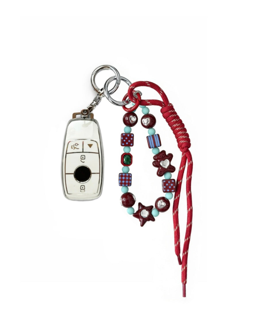 White Mercedes Benz car key fob cover case with pastel plaid heart design, paired with a handmade beaded keychain featuring cute patterned beads, and a red braided cord on a white background.